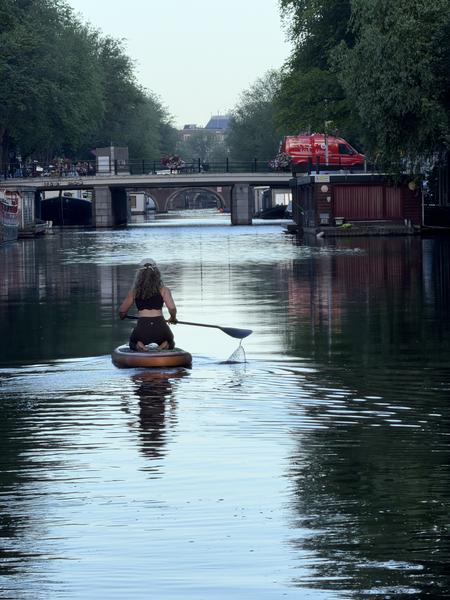 A person kneels on a paddleboard, paddling along the tranquil Nieuwe Prinsengracht canal in Weesperbuurt, Amsterdam, with lush green trees lining the banks and a bridge in the distance